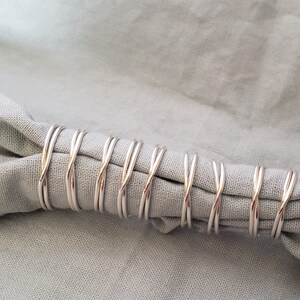 May include: Close-up of a light grey linen napkin held by several silver napkin rings. The napkin rings have a twisted design. The napkin is folded and draped, creating soft folds and shadows.