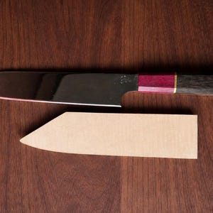 May include: A stainless steel chef's knife with a dark wood handle accented with red and gold. The knife is paired with a light brown sheath, all set against a dark wood grain surface. A high-quality kitchen tool.