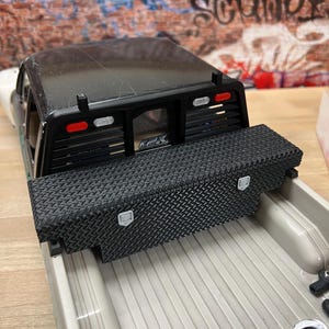 Puede incluir: Una caja de camión negra con una caja de herramientas de chapa de diamante en la parte trasera. La caja de herramientas tiene dos cierres y está hecha de plástico negro. La caja del camión está hecha de plástico gris y tiene una superficie texturizada.