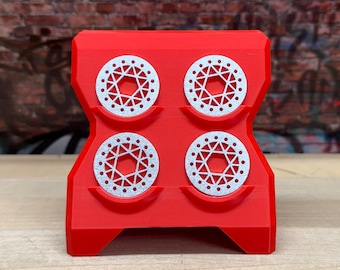 Op de afbeelding: Een rode 3D-geprinte houder met vier zilverkleurige, cirkelvormige objecten. Elk object heeft een geometrisch ontwerp met een reeks kleine gaatjes. De houder staat op een houten oppervlak met een bakstenen muur op de achtergrond.