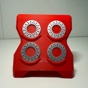 以下が含まれることがあります： 赤色の3Dプリントホルダー。4つの円形、銀色の金属ディスクが付いています。各ディスクには周囲に小さな穴が並んでいます。ホルダーは、幅広のベースと湾曲した上部を備えた、ユニークな幾何学的形状をしています。