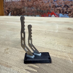 Puede incluir: Un par de objetos plateados con forma de palanca y empuñaduras texturizadas, montados sobre una base rectangular negra. Los objetos están inclinados hacia arriba. El fondo presenta una superficie de madera y un muro de ladrillo con grafitis.