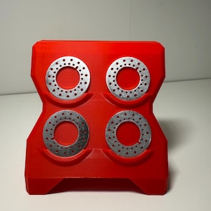 Op de afbeelding: Een helderrode houder met vier zilverkleurige metalen schijven, elk met kleine gaatjes. De houder heeft een geometrisch ontwerp en staat tegen een effen witte achtergrond.
