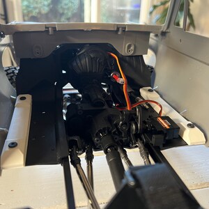 May include: A close-up view of the interior of a gray and white model car. The car's engine and chassis are visible, along with a black servo motor and orange and white wires.