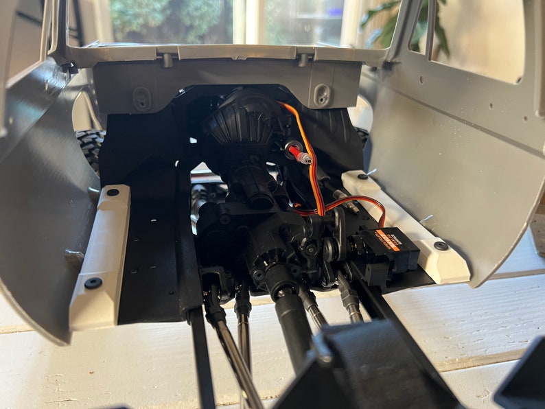 May include: A close-up view of the interior of a gray and white model car. The car's engine and chassis are visible, along with a black servo motor and orange and white wires.