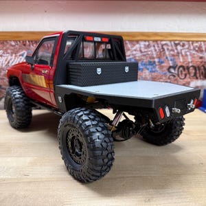 Könnte beinhalten: Ein rotes ferngesteuertes Pickup-Modell mit Pritsche, schwarzer Werkzeugbox mit Riffelblech-Optik, schwarzem Überrollbügel und großen Geländereifen. Das Fahrzeug verfügt über eine detaillierte Heckstoßstange mit integrierten roten und weißen Rückleuchten.