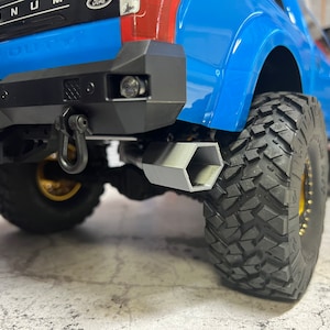 May include: A blue pickup truck with a black rear bumper and a silver tow hitch. The truck has a black exhaust pipe and large black tires.