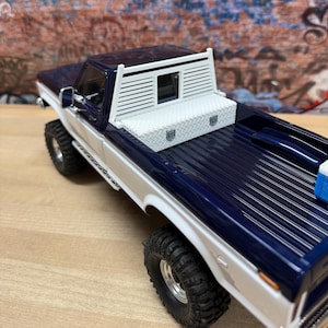 May include: A dark blue and white scale model pickup truck featuring a white headache rack and a white diamond plate tool box mounted in the truck bed. The vehicle has large black off-road tires, chrome side mirrors, and a blue cooler in the back.