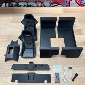 May include: A set of black 3D printed car interior parts, including two seats, a dashboard, a center console, and various other components. The parts are laid out on a wooden surface.