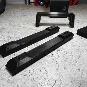 May include: Three black plastic pieces for a 3D printer. Two long pieces with holes and a shorter piece with a rectangular shape.