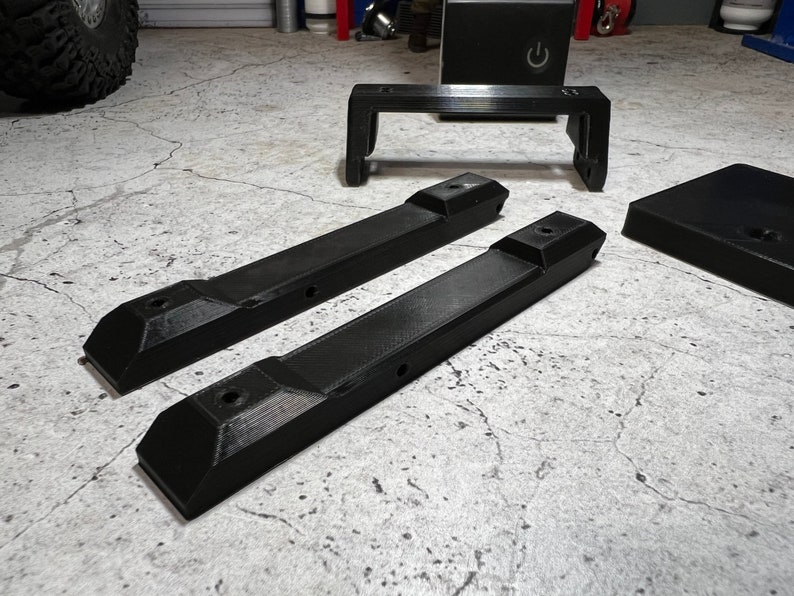 May include: Three black plastic pieces for a 3D printer. Two long pieces with holes and a shorter piece with a rectangular shape.