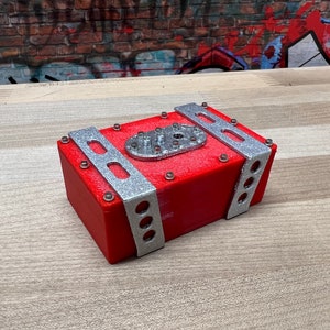 May include: A red fuel cell with silver metal brackets and rivets. The fuel cell has a round opening on the top.