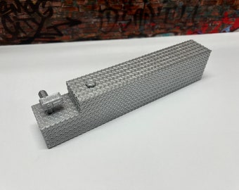 May include: A silver metal model of a fuel tank with a diamond plate texture. The tank has a small, round cap on top and a metal bracket on the side.