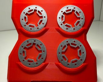 May include: A red 3D-printed holder with four silver metal discs. Each disc has a star-like design with multiple holes. The holder has a unique, geometric shape, and the discs are evenly spaced within it.