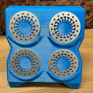 Könnte beinhalten: Ein blauer 3D-gedruckter Halter mit vier silbernen Untersetzern. Jeder Untersetzer hat ein zentrales Blitzdesign, das von kleinen Löchern umgeben ist. Der Halter ist so konzipiert, dass er die Untersetzer aufnimmt und eine moderne, geometrische Ästhetik zeigt.