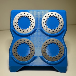 Puede incluir: Un soporte azul con cuatro discos de freno plateados. Cada disco es circular con un patrón de pequeños agujeros alrededor del borde. El soporte está diseñado para guardar y organizar los discos de forma segura.