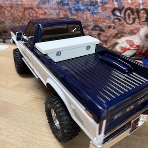 May include: A scale model pickup truck featuring a two-tone dark blue and white exterior paint scheme. The truck bed includes a white rectangular storage box mounted behind the cab, black off-road tires, and chrome-colored trim details.