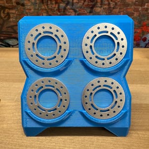 Puede incluir: Un soporte azul impreso en 3D con cuatro discos metálicos circulares plateados. Cada disco tiene un agujero central y múltiples agujeros más pequeños alrededor del perímetro. El soporte es rectangular con cortes curvos.