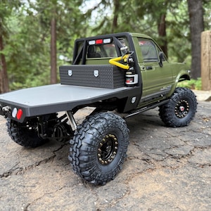 May include: A detailed model of a green pickup truck with a flatbed. The truck features large, black off-road tyres, a black toolbox, and a roll bar with lights. The model is set against a backdrop of trees and a rocky surface.