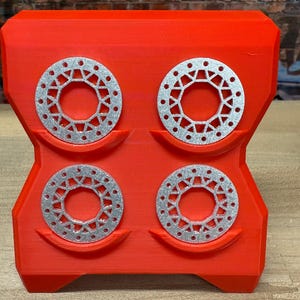 Op de afbeelding: Een felrode 3D-geprinte organizer met vier zilverkleurige, cirkelvormige objecten met een patroon. De organizer heeft een unieke, hoekige vorm en is ontworpen om de cirkelvormige objecten op hun plaats te houden.