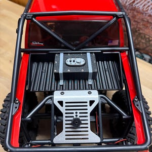 以下が含まれることがあります： ロールケージ付きの赤と黒のラジコンカー。トラックは黒い荷台と背面に銀色の金属板が付いています。タイヤは黒で、トラックの背面にはアメリカ国旗のステッカーが付いています。