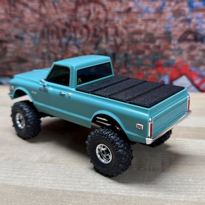Peut inclure: Un modèle réduit de camionnette Chevrolet turquoise avec de gros pneus tout-terrain noirs. Le camion a une bâche de lit noire et des garnitures chromées. L'arrière-plan présente un mur de briques avec des graffitis.