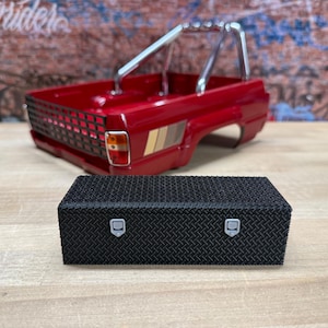 May include: A black rectangular diamond plate toolbox with two silver latches positioned in front of a red toy truck bed. The truck bed features a chrome roll bar, a black grid tailgate, and a side decal with tan, brown, and white stripes.