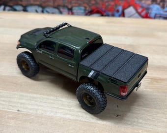 May include: A die-cast olive green pickup truck model featuring a black textured folding tonneau cover, off-road tires with bronze wheels, a roof-mounted light bar, and TRD Off-Road decals on the rear quarter panels with a small license plate.