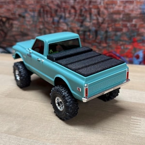 Peut inclure: Un modèle de pick-up Chevrolet turquoise avec de gros pneus tout-terrain noirs. La benne du camion a une couverture noire. Le mot "CHEVROLET" est visible sur le hayon. Le camion est posé contre un mur de briques.