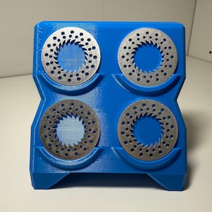 May include: A blue 3D-printed holder containing four silver brake discs. Each disc has a series of small holes around the edge. The holder has an angular design. The item is on a white surface.