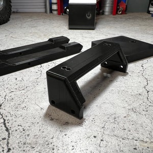 May include: Three black 3D printed parts for a model car. The parts are a bumper, a chassis, and a base. The parts are on a gray concrete surface.