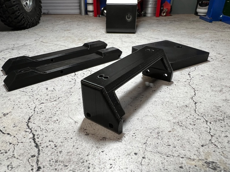 May include: Three black 3D printed parts for a model car. The parts are a bumper, a chassis, and a base. The parts are on a gray concrete surface.