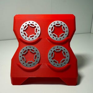 May include: A red plastic display stand holding four silver metal discs. Each disc features a distinctive star-shaped design with multiple holes. The stand is designed to securely hold the discs, highlighting their intricate patterns and metallic finish.