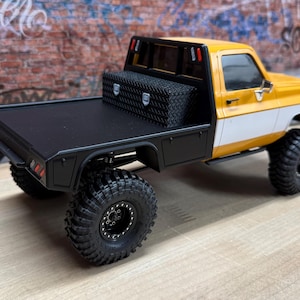 May include: A yellow and white toy truck with a black flatbed and large, black off-road tires. A black toolbox is mounted on the flatbed. The truck has a detailed design, including side mirrors and a black roll bar.