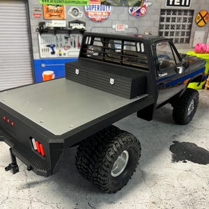 May include: Black pickup truck with a flatbed and a black metal toolbox in the bed. The truck has a black metal grill and a black metal bumper. The truck is sitting on a gray concrete surface.