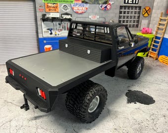 May include: Black pickup truck with a flatbed and a black metal toolbox in the bed. The truck has a black metal grill and a black metal bumper. The truck is sitting on a gray concrete surface.