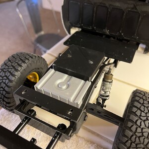 May include: A black and silver toy truck chassis with a black fuel tank and black wheels. The truck has a black metal frame and a silver plastic fuel tank. The truck is sitting on a white surface.
