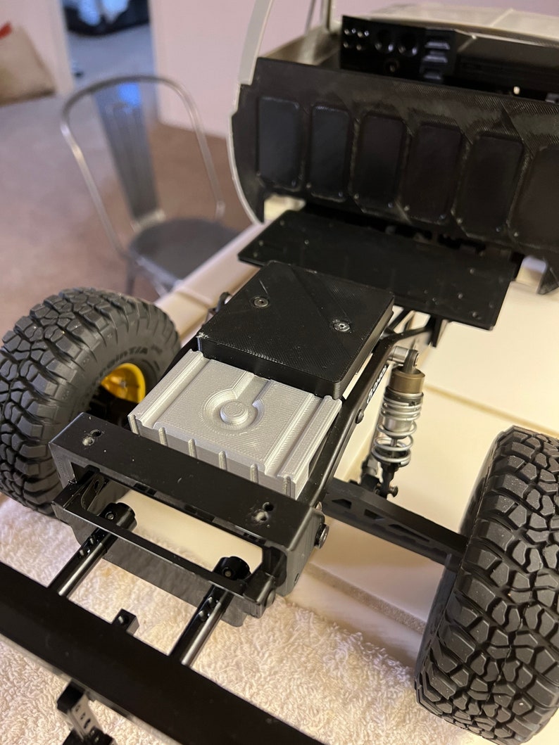 May include: A black and silver toy truck chassis with a black fuel tank and black wheels. The truck has a black metal frame and a silver plastic fuel tank. The truck is sitting on a white surface.