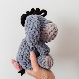 May include: A handmade crochet plush toy of a donkey character with a light blue body, a lighter gray muzzle, a dark black mane, and a small pink tail. The stuffed animal features a soft, textured yarn construction with long, drooping ears.
