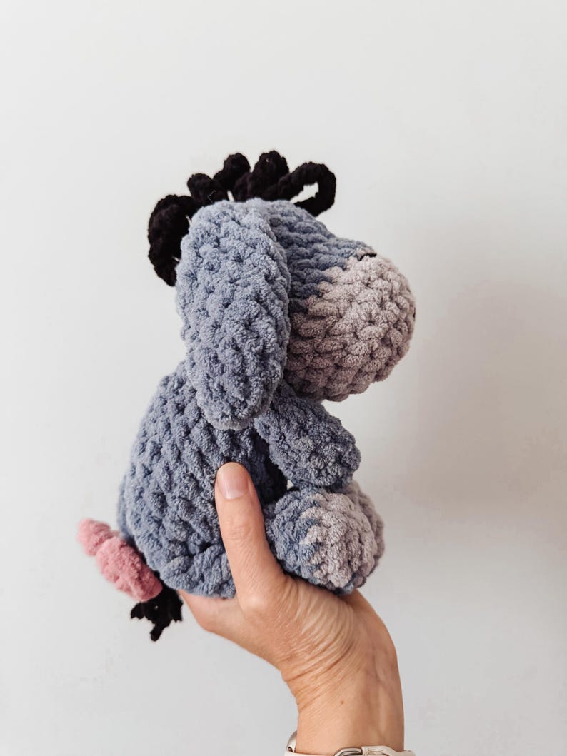 May include: A handmade crochet plush toy of a donkey character with a light blue body, a lighter gray muzzle, a dark black mane, and a small pink tail. The stuffed animal features a soft, textured yarn construction with long, drooping ears.