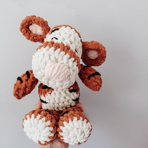 May include: A plush tiger toy crafted from chunky, textured yarn in shades of burnt orange, cream, and black. The stuffed animal features large rounded ears, a prominent snout, and striped markings across its body, limbs, and feet.