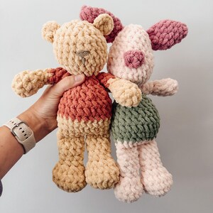 May include: Two handmade crochet plush toys made from chunky yarn. One is a tan bear wearing a red shirt, and the other is a light pink pig with dark pink ears and a green shirt. Both toys feature a soft, textured stitch pattern and rounded limbs.