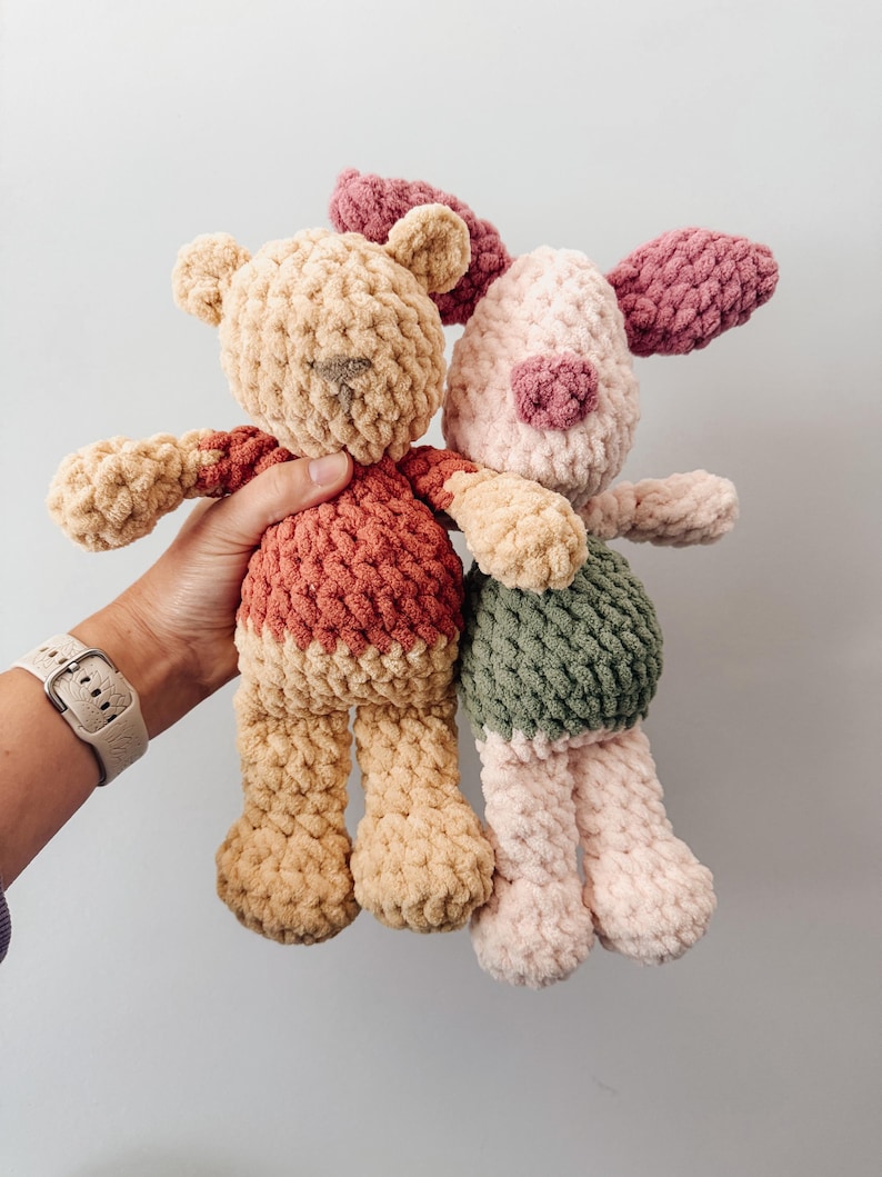 May include: Two handmade crochet plush toys made from chunky yarn. One is a tan bear wearing a red shirt, and the other is a light pink pig with dark pink ears and a green shirt. Both toys feature a soft, textured stitch pattern and rounded limbs.