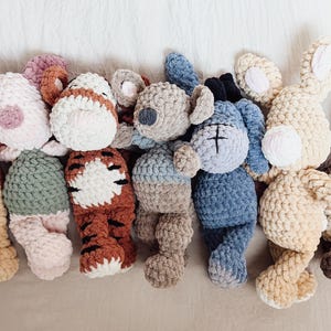 May include: A collection of seven handmade crochet plush toys featuring various animal designs. The set includes a tan bear, a pink and green pig, a tiger with brown stripes, a gray koala, a blue donkey, a tan rabbit, and a brown and white owl.