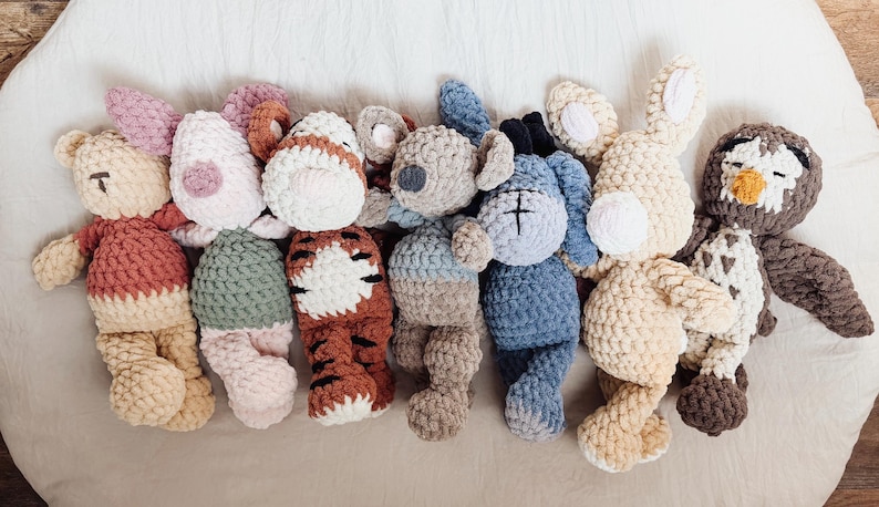 May include: A collection of seven handmade crochet plush toys featuring various animal designs. The set includes a tan bear, a pink and green pig, a tiger with brown stripes, a gray koala, a blue donkey, a tan rabbit, and a brown and white owl.
