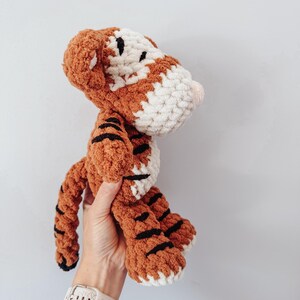 May include: A crocheted tiger plush toy made with thick, textured yarn in shades of orange, white, and black. The stuffed animal features a rounded head, small ears, a long tail, and distinct black stripes across its body and limbs.