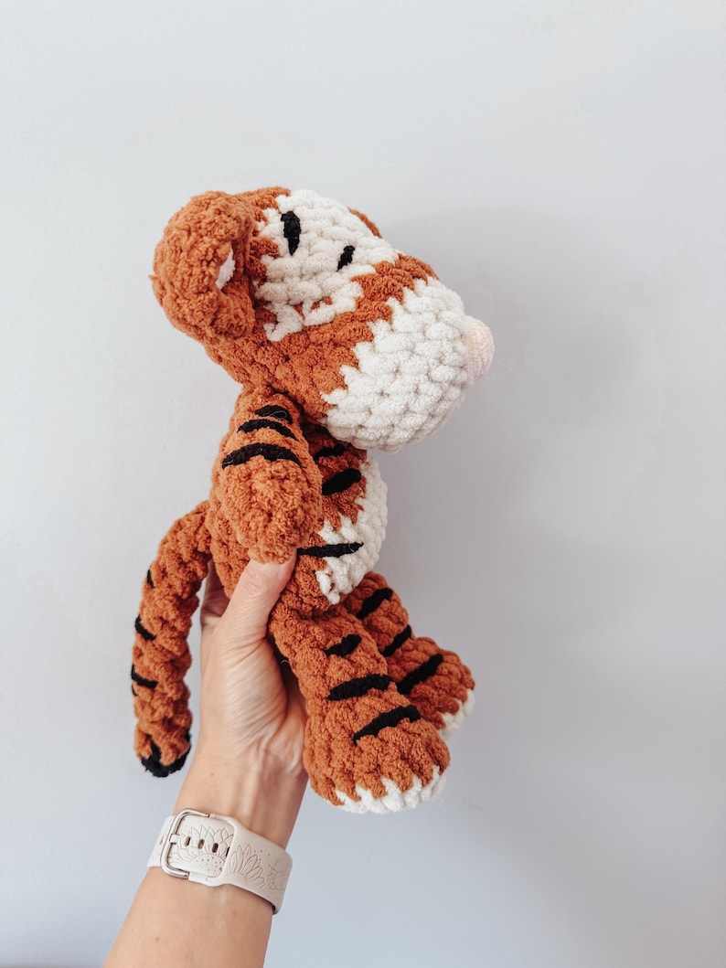 May include: A crocheted tiger plush toy made with thick, textured yarn in shades of orange, white, and black. The stuffed animal features a rounded head, small ears, a long tail, and distinct black stripes across its body and limbs.