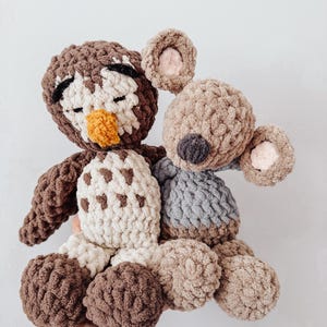 Puede incluir: Un par de peluches de ganchillo que consiste en un búho marrón y crema con pico amarillo y ojos cerrados, y un koala marrón claro con nariz oscura y orejas forradas de rosa, ambos hechos de hilo grueso y texturizado de estilo suave.