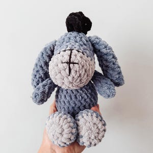 May include: A plush donkey toy made of blue and light gray yarn with a soft, textured finish. The character features long, drooping ears, a black tuft of hair on top, and a stitched cross detail on its snout. The body is round and compact.