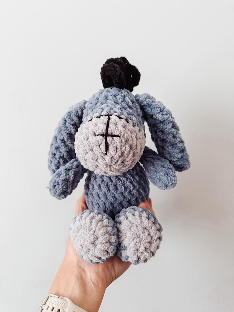 May include: A plush donkey toy made of blue and light gray yarn with a soft, textured finish. The character features long, drooping ears, a black tuft of hair on top, and a stitched cross detail on its snout. The body is round and compact.